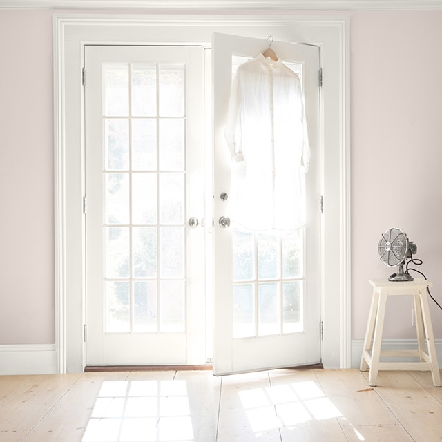 White-painted French doors opening in a pinkish beige room. A fan sits on a white stool, and a white button up is hanging on a door.