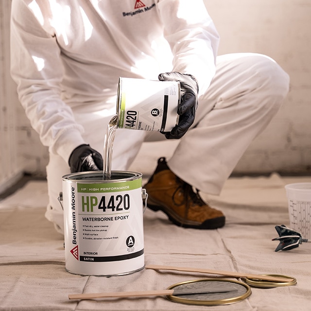 A person combining two components of HP4420 Waterborne Epoxy, pouring a small can into a larger gallon container.