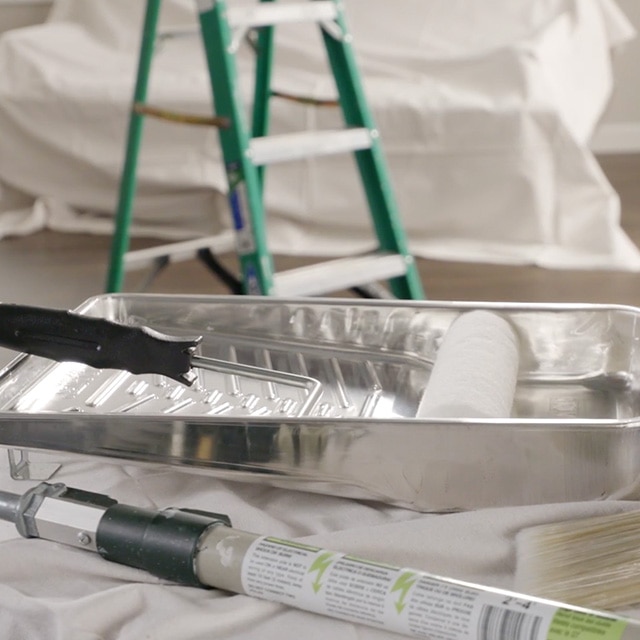 Un rouleau dans un bac de peinture et différents outils de peinture sur une toile de protection devant une échelle verte.