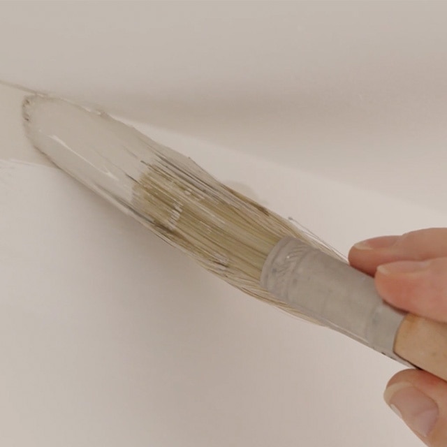 Gros plan d’une personne qui découpe à la peinture blanche la ligne entre le haut d’un mur et le plafond.
