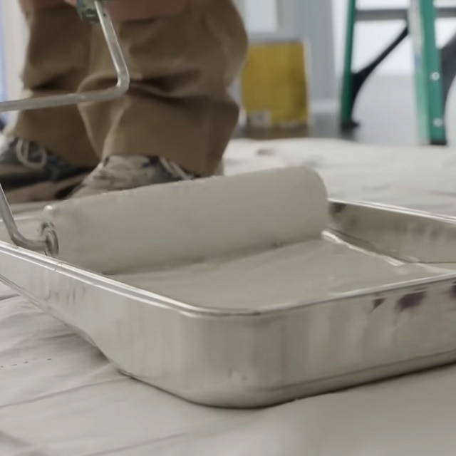 Gros plan d’une personne qui trempe son rouleau en le roulant le long de la partie surélevée du bac rempli de peinture blanche.