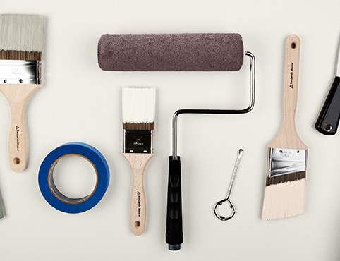 A collection of painting supplies, including paintbrushes, a paint roller, painter’s tape, a paint tray containing brown paint, and more.