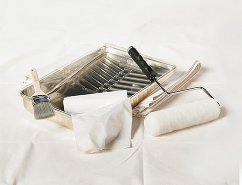 Painting supplies including a metal paint tray, an angled sash brush, a paint roller and a plastic drop cloth.