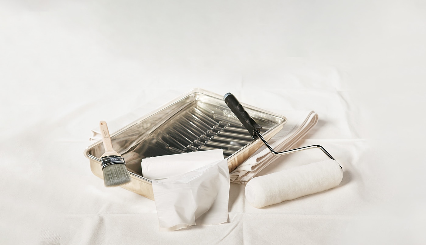 Painting supplies including a metal paint tray, an angled sash brush, a paint roller and a plastic drop cloth.