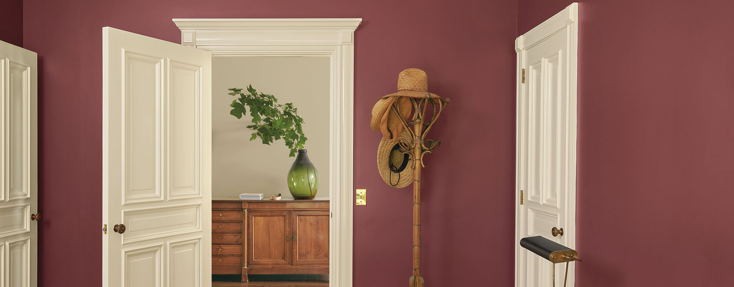 Una bonita habitación pintada de rojo con marcos y puertas de color beige, una cómoda cama con sábanas blancas y una almohada naranja, y un perchero junto a una puerta abierta que da a un pasillo pintado de gris.