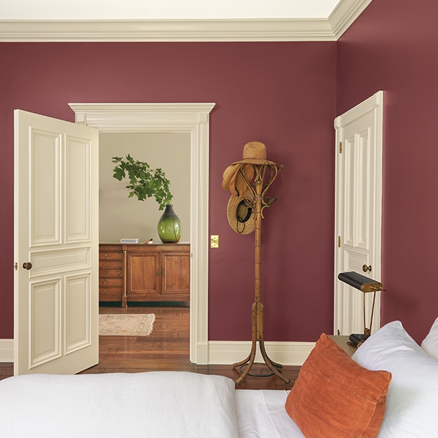 Una bonita habitación pintada de rojo con marcos y puertas de color beige, una cómoda cama con sábanas blancas y una almohada naranja, y un perchero junto a una puerta abierta que da a un pasillo pintado de gris.