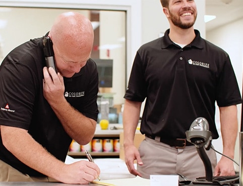 Two Benjamin Moore paint retailers wearing black shirts behind a counter, one of them talking on the phone and taking notes.