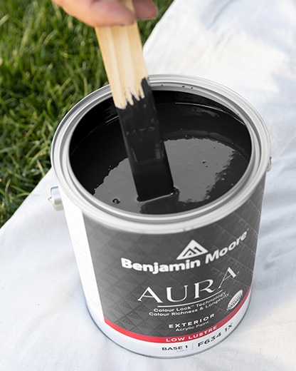 An open can of black AURA® exterior paint being stirred, on top of a tarp.