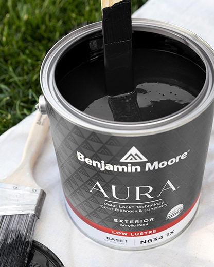 An open gallon of black AURA® exterior paint being stirred, on top of a tarp.