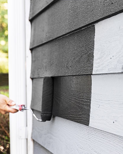 A person using a paint roller to apply black AURA® Exterior paint onto siding, with a white downspout.