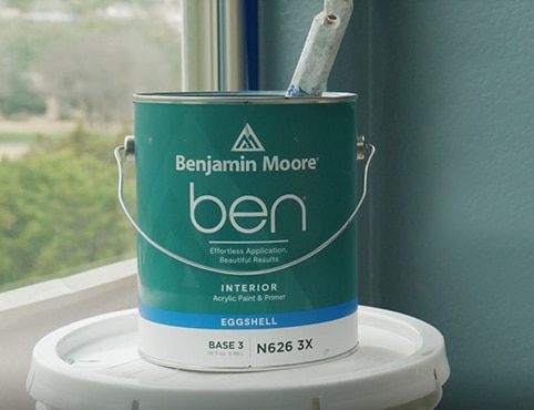 An open can of ben Interior paint on top of a white bucket, in front of a window and blue painted wall.