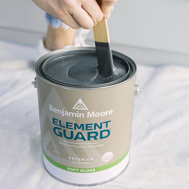 A person stirring an open gallon of blue Element Guard® exterior paint on top of a white floor tarp.