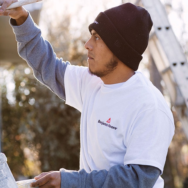 A painting contractor wearing a black cap and white and blue shirts, holding a can of Element Guard®, painting the exterior of a home with a brush, and standing next to a tall ladder.