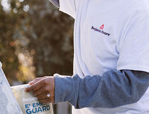 A painting contractor wearing white and blue shirts, holding a can of Element Guard®, painting the exterior of a home with a brush, and standing next to a tall ladder.