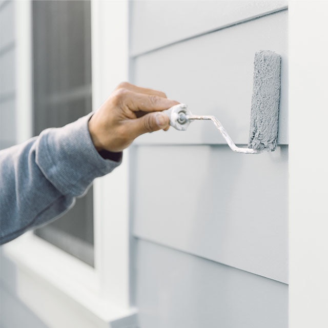 Une personne applique au rouleau une couche de peinture gris sur un parement extérieur.