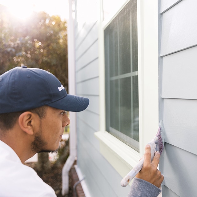 Un peintre professionnel portant une tuque bleue applique de la peinture gris pâle sur le parement d’une maison, près du rebord d’une fenêtre blanche.