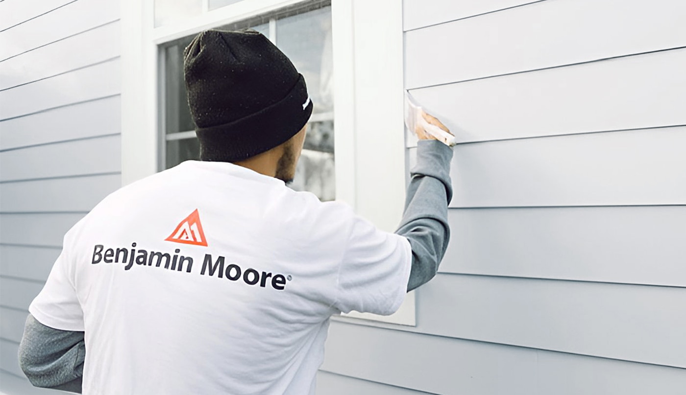 Un peintre professionnel portant une tuque bleue applique de la peinture gris pâle sur le parement d’une maison, près du rebord d’une fenêtre blanche.