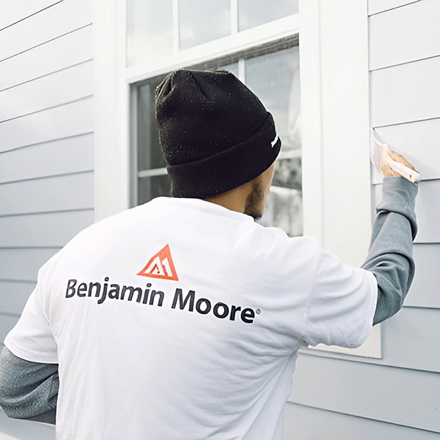 Un peintre professionnel portant une tuque bleue applique de la peinture gris pâle sur le parement d’une maison, près du rebord d’une fenêtre blanche.