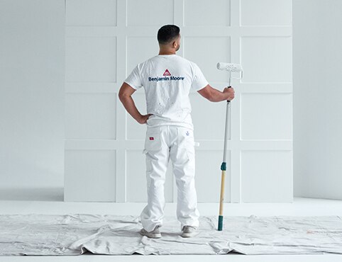  Benjamin Moore professional painting contractor standing in a white-painted room, facing a panelled section of wall, and holding a paint roller.