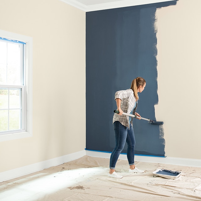 A woman paints dark blue paint over a beige wall using a roller surrounded by drop cloths on floor and table.