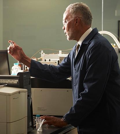 Dans un laboratoire gris, un chimiste portant des lunettes de protection étanches et un sarrau noir tient une fiole devant des appareils de laboratoire.