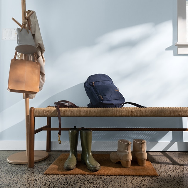 A sunlit, blue-gray painted hallway wall with light gray trim, a wood bench with boots underneath, and a coatrack with hanging bags.