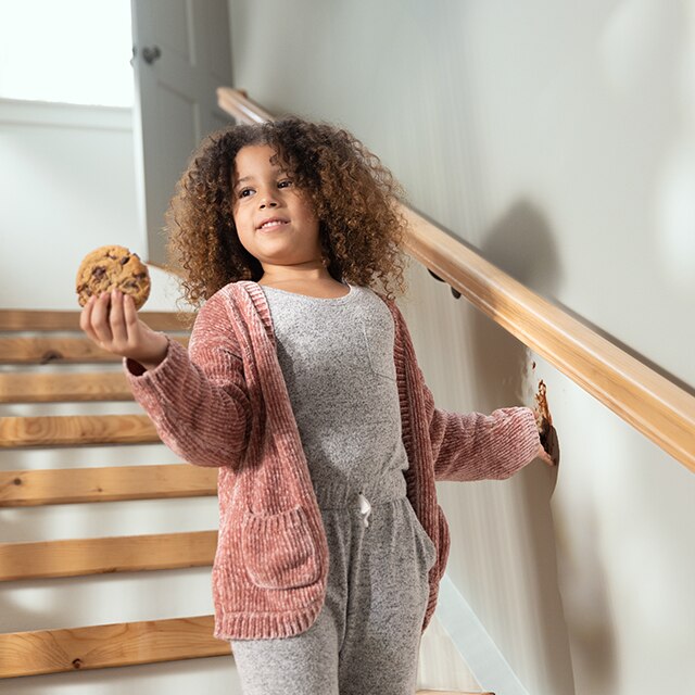 A young girl standing on a staircase with a cookie in one hand and touching a light gray-painted wall with her other hand.