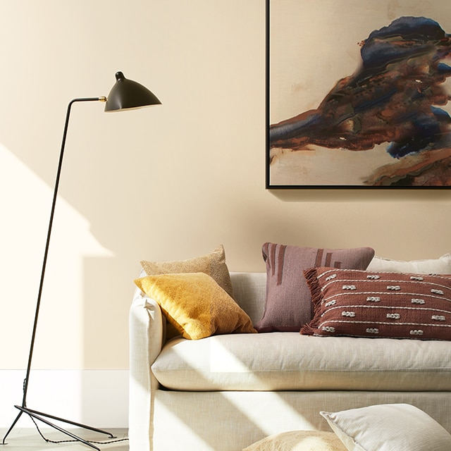 Un canapé décontracté blanc cassé avec des coussins jaunes et rouges placé contre un mur peint en beige avec des tons nuancés de pêche, une œuvre d’art contemporaine et un lampadaire noir.