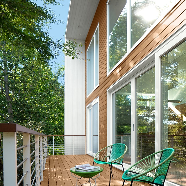 Two modern green rattan chairs and a table sit atop a deck with light brown wood flooring and white railings, extending from a home exterior sided with light brown wood, white trim, and floor-to-ceiling windows.