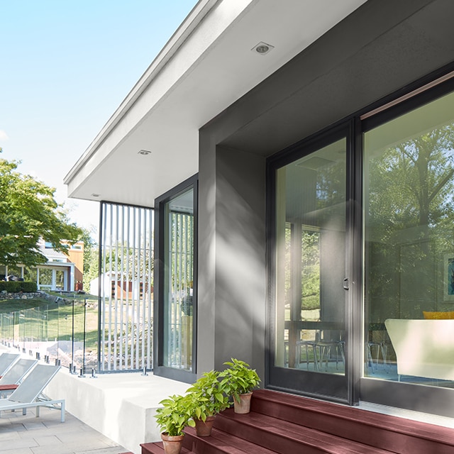 Three green plants sit on red stained wood steps leading to a red stained deck outside of a house with charcoal gray siding and sliding glass doors.