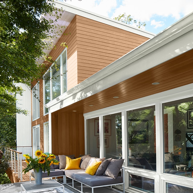 A gray deck with a blue outdoor couch and yellow pillows, backs up to a one-story enclosed porch with brown wood siding and white painted trim, attached to a two-story home with light brown siding and white trim.