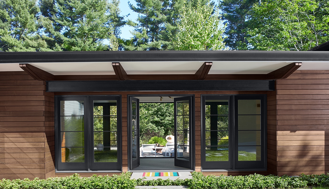 A modern one-story home with brown wood siding and black trim features floor-to-ceiling windows and open French doors looking through to the backyard.