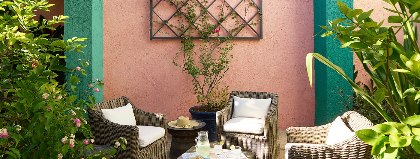 A pretty coral and green painted wall with trellis plays backdrop to an inviting outdoor seating area with wicker chairs and table, flanked by tropical plants and flowers.