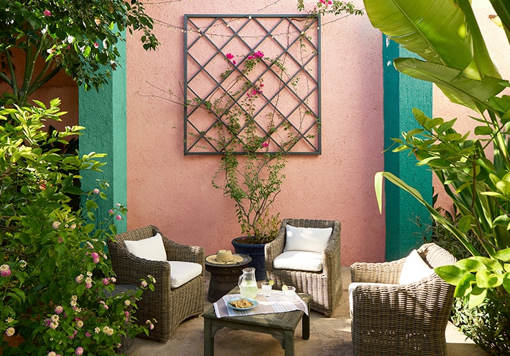 A pretty coral and green painted wall with trellis plays backdrop to an inviting outdoor seating area with wicker chairs and table, flanked by tropical plants and flowers.