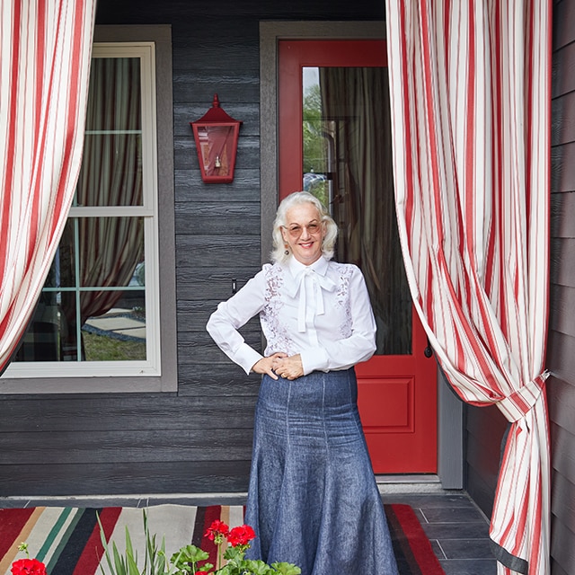 Interior designer Alessandra Branca stands on the front porch of the bungalow she designed, framed in red and white striped curtains with a charcoal gray home exterior and bold, red-painted front door behind her.