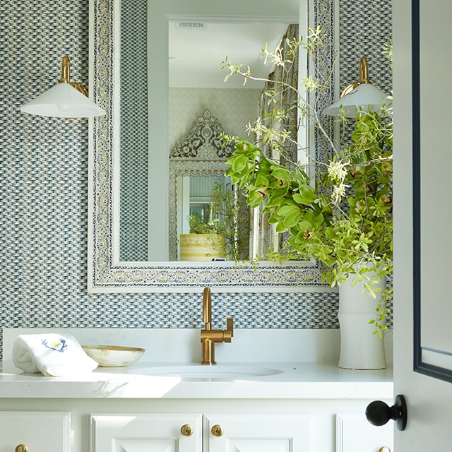 Une salle de bains présentant une vanité peinte en blanc, du papier peint bleu et blanc, des accessoires en cuivre et un bouquet de fleurs sauvages dans un vase blanc sur un comptoir blanc.