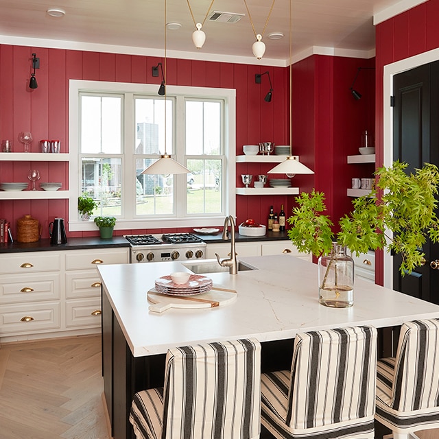 Une cuisine arborant des murs peints en rouge, des moulures blanches, un îlot de cuisine surmonté d’un comptoir blanc, des comptoirs noirs et des portes peintes en noir.