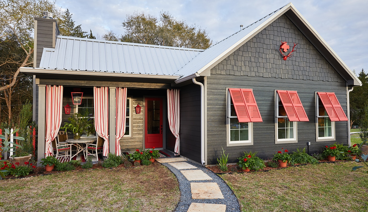 The exterior of a luxury cottage with dark, gray-painted siding, red shutters and front door, and a front porch accented with red and white striped curtains.