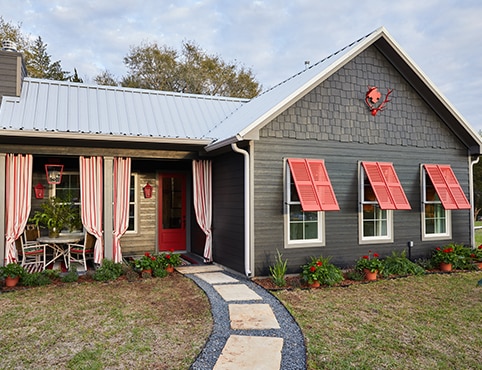 The exterior of a luxury cottage with dark, gray-painted siding, red shutters and front door, and a front porch accented with red and white striped curtains.