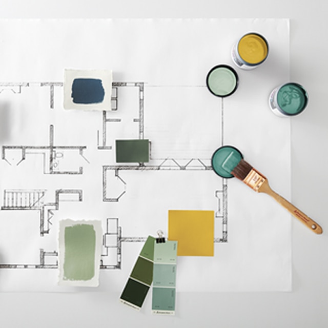A bird’s eye view sees a variety of Benjamin Moore color swatches, color chips and 8 oz. color samples, plus a wooden paint mixer and a paint brush, atop a printed floor plan of a building.