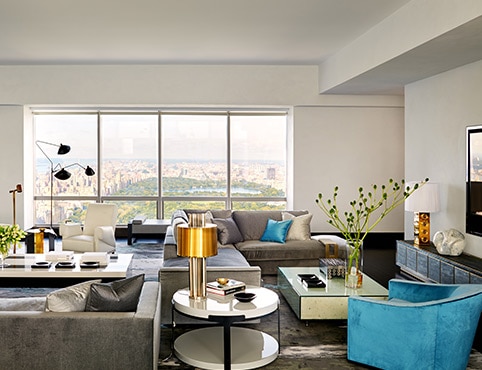 A Manhattan apartment living room with white-painted walls, gray sectional sofas, blue armchair, and a glass cocktail table.