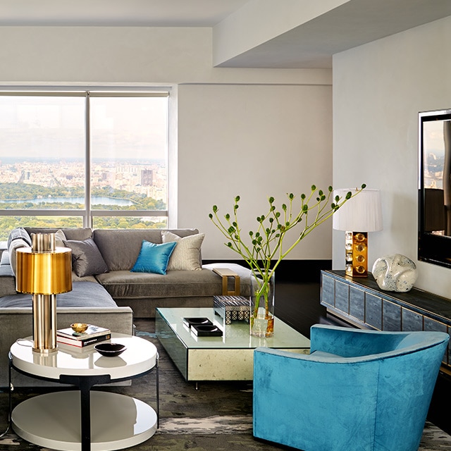 A Manhattan apartment living room with white-painted walls, gray sectional sofas, blue armchair, and a glass cocktail table.
