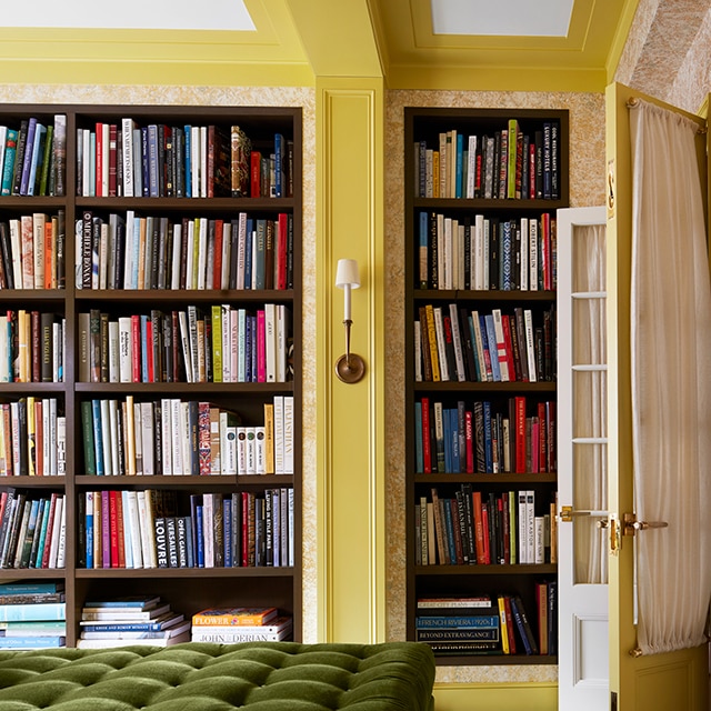 Une magnifique bibliothèque avec des murs et moulures jaunes, un plafond blanc aux poutres jaunes, des étagères pleine longueur et un large pouf touffeté vert.