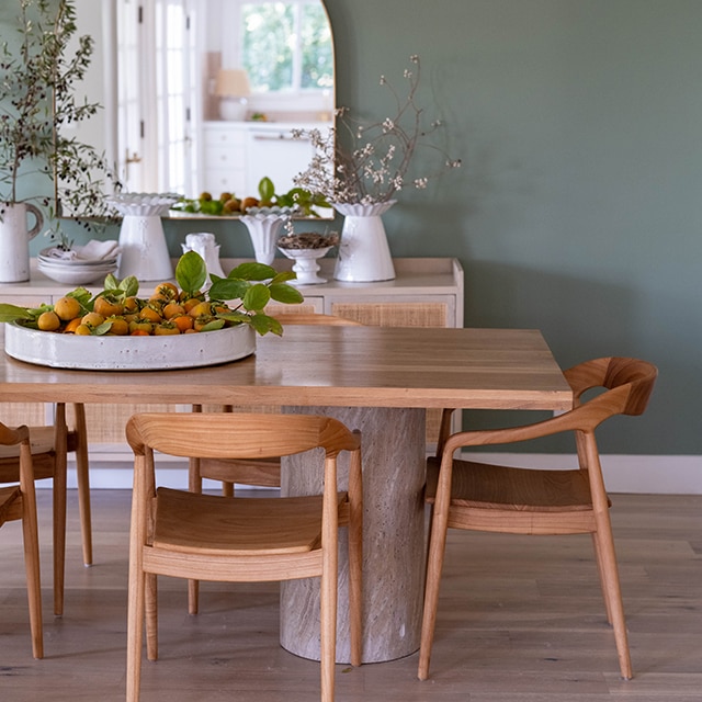 Pretty wood modern dining table, chairs and sideboard in front of a green-painted wall, with a large mirror, and white hanging light.