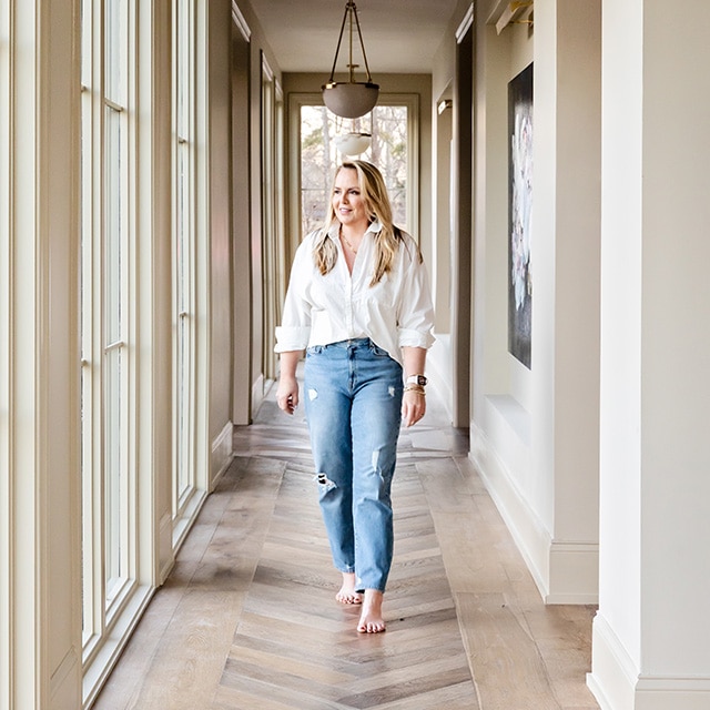 Lori Paranjape walks down a beige hallway with wood flooring and floor-to-ceiling windows.