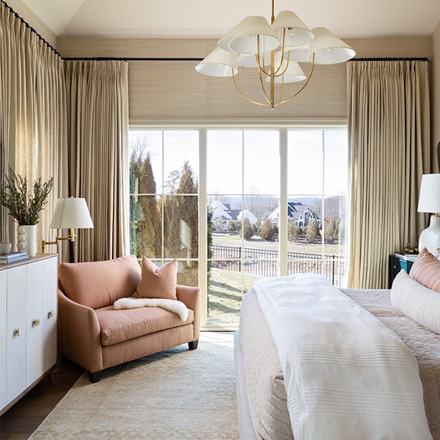 A bedroom with a white painted ceiling, golden curtains lining both the walls and windows, and a beige carpet over a dark brown floor.