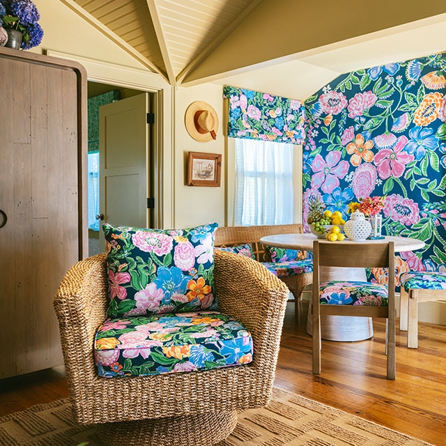 Un comedor y sala de estar pintados en color tostado oscuro, con muebles de junco y madera, papel pintado floral colorido, y cojines y almohadas florales a juego.