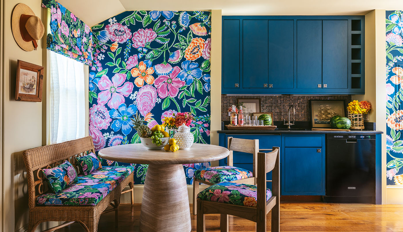 Un comedor y cocina alegres, con papel pintado floral vibrante y cojines de asiento a juego, paredes y techo pintados en color tostado oscuro, y gabinetes azul oscuro.