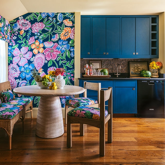 Un comedor y cocina alegres, con papel pintado floral vibrante y cojines de asiento a juego, paredes y techo pintados en color tostado oscuro, y gabinetes azul oscuro.