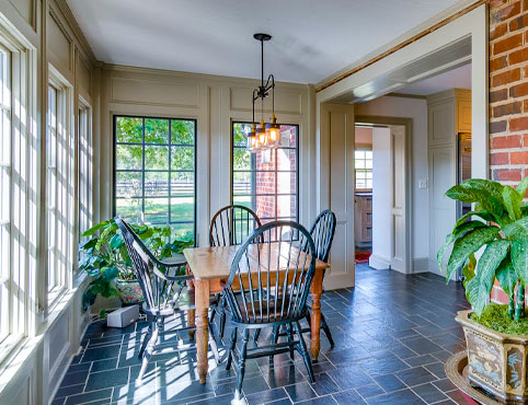 Sunroom with one brick wall and three white-painted walls with large windows, featuring a wooden table and chairs and a variety of plants.